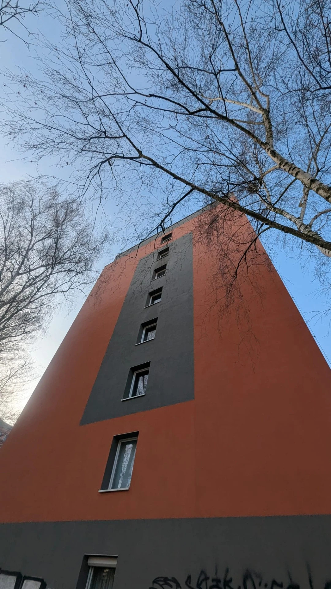 Červenobílá výšková budova s okny, pohled zdola proti modré obloze a holým větvím stromů.