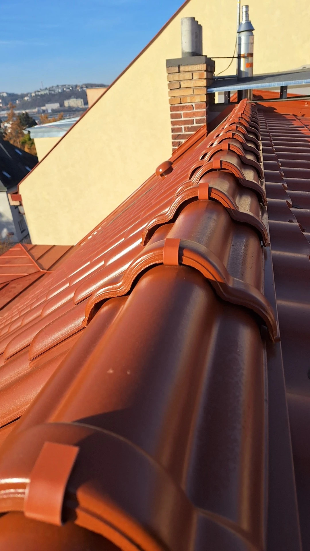 Detail červené střechy s taškami, komínem a kovovým odvětráním na pozadí modré oblohy.