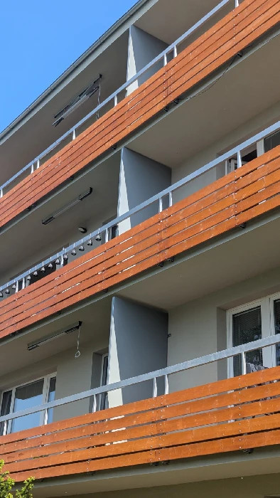 Moderní bytový dům s dřevěnými balkony, bílými zábradlími a otevřenými okny pod modrou oblohou.