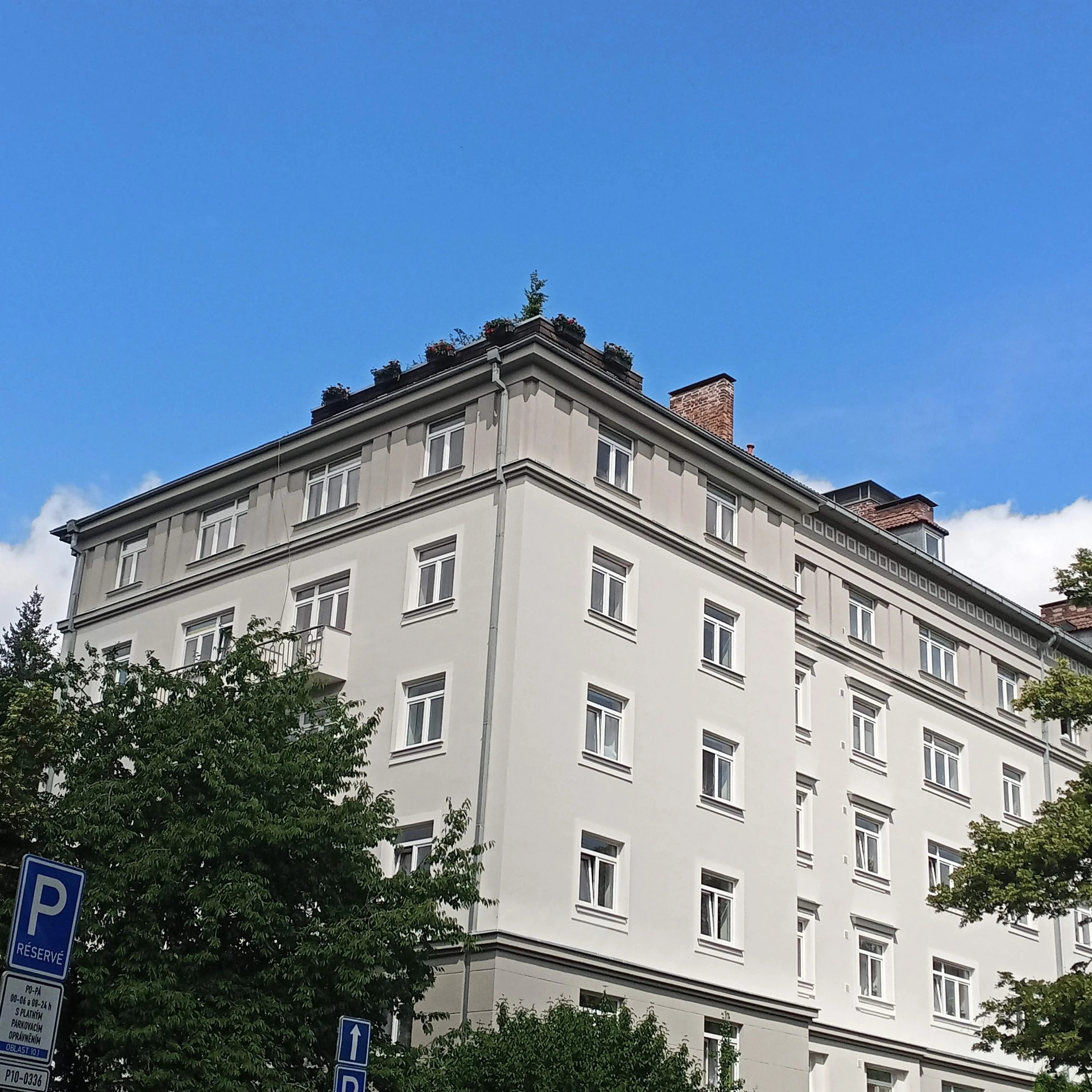 Bílá bytová budova s řadami oken, šikmou střechou a komínem, na pozadí modrá obloha.