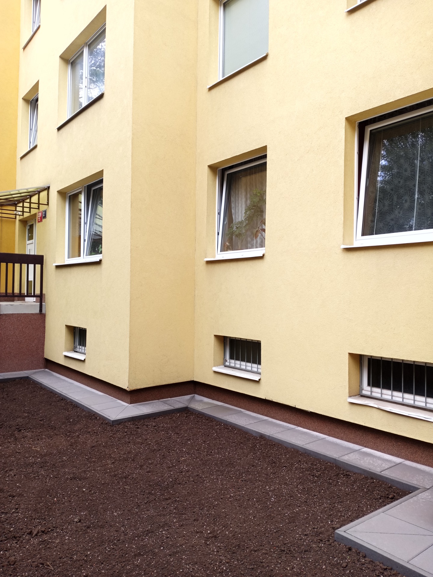 Žlutá fasáda bytového domu s několika okny, parapety a přilehlou dlážděnou chodbou u vchodu.