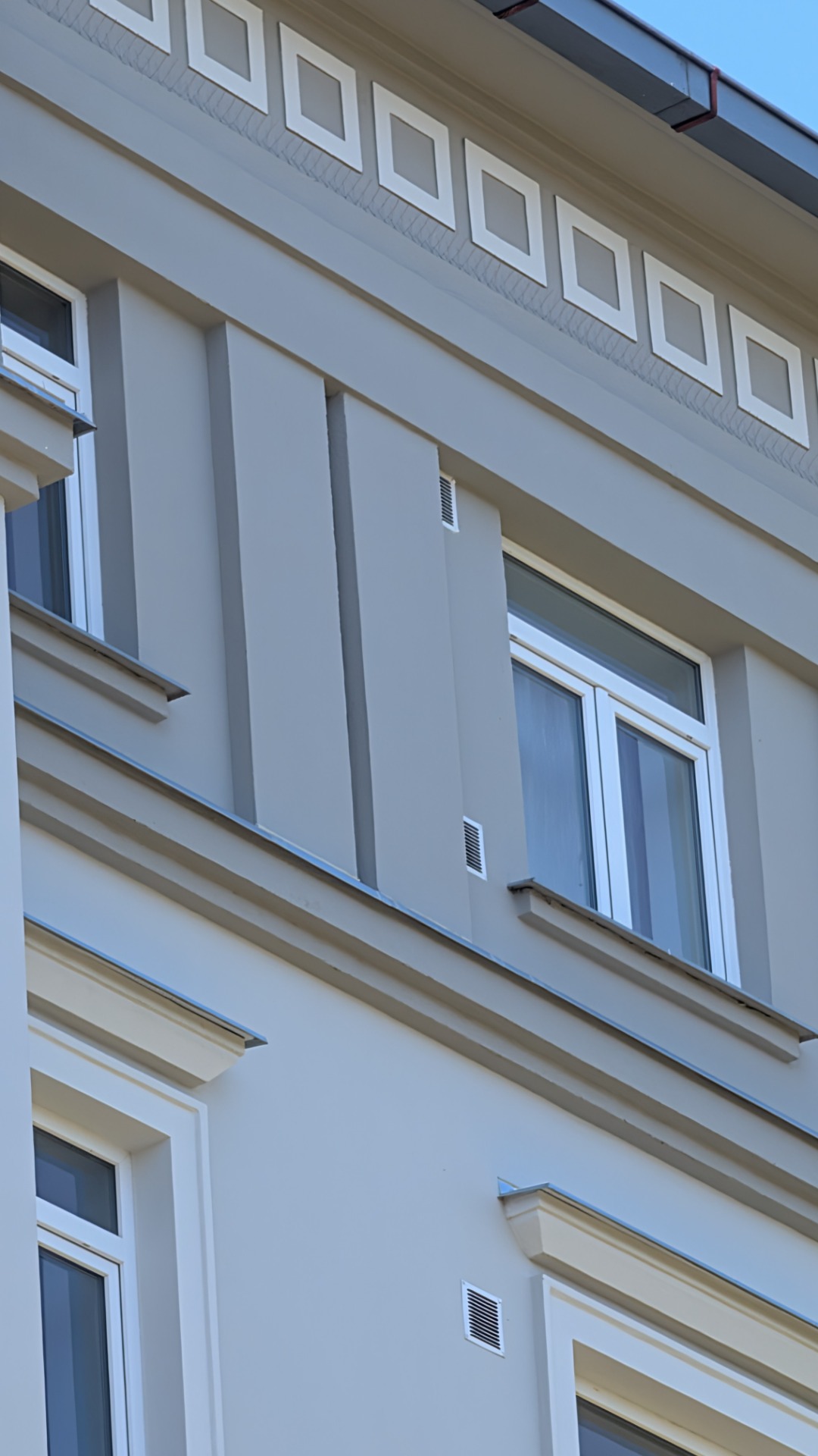 Bílá bytová budova s řadami oken, šikmou střechou a komínem, na pozadí modrá obloha.